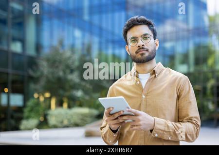 Confident hispanic man wearing glasses and casual clothes using digital tablet outdoors near modern office building. Urban office background with greenery and glass architecture. Stock Photo