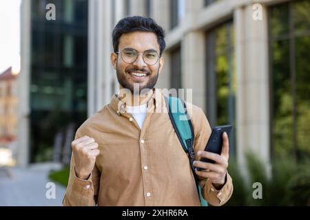 Hispanic student holding smartphone looking happy and celebrating success outside campus building. Young man with backpack showing excitement. Concept of achievement, education, and student life. Stock Photo