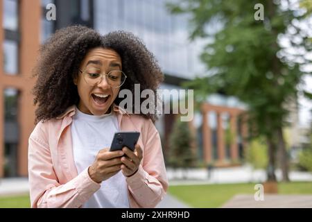 Joyful woman student with curly hair holding smartphone outdoors on campus. Young woman using mobile phone, looking pleasantly surprised and happy. Urban outdoor setting. Stock Photo