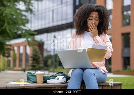 Woman student looking worried while reading letter on laptop outdoors at university campus. Concept of education, stress, university life, bad news, financial issues. Stock Photo