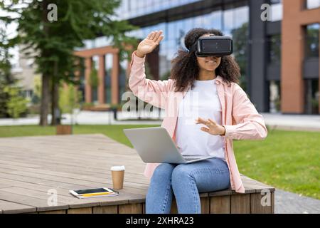 Woman student using VR headset outdoors with laptop and coffee cup nearby. Enjoying technology in open-air environment. Smiling and engaging in virtual experience outside modern building Stock Photo