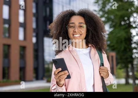 Happy woman student with glasses holding smartphone outdoors in urban setting. Youthful, cheerful expression showcasing relaxation and digital connectivity near modern building. Concept of education, technology, and casual lifestyle. Stock Photo