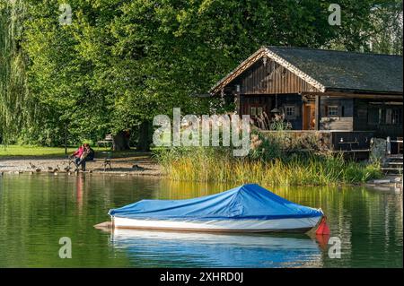 Wooden hut with common reed (Phragmites australis), covered boat, two woman on a park bench, shore of Lake Schliersee, spa garden Markt Schliersee Stock Photo