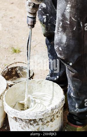 Paddle mixer for cement adhesive mortar connects to electric drill for decorating and building jobs, construction worker mixes tile glue mortar in buc Stock Photo