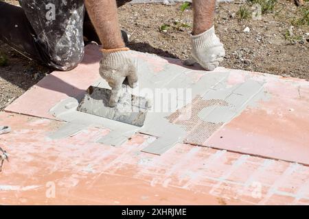 Construction worker uses steel trowel to apply adhesive cement mortar to flexible faux brick panel for exterior wall of house. Stock Photo