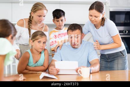 Frustrated man sits in kitchen next to family and reads unpleasant news in letter. Stock Photo