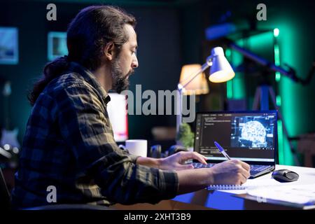 Man at desk using laptop for programming and data analysis. Male programmer working at home, using his notepad while efficiently managing information technology and data center on digital device. Stock Photo