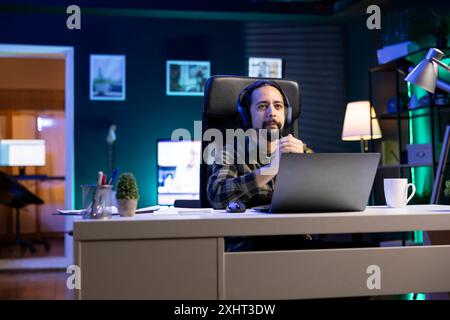 A male college student is using a digital laptop to browse the internet while sporting wireless headphones. Young man using a portable computer at home and a headset to listen to music. Stock Photo