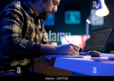 Diligent man multitasking, writing notes while reviewing forex chart on his laptop. Male freelancer working from home, using his notebook and checking status of stock market trend values on device. Stock Photo
