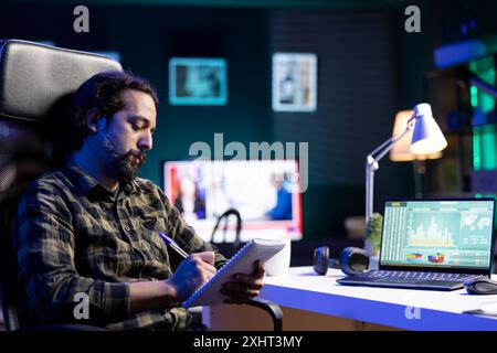 Busy youthful man working at home, multitasking on his laptop, planning, reviewing documents, and researching online. Male freelancer writing on notebook while sitting at his desk. Stock Photo