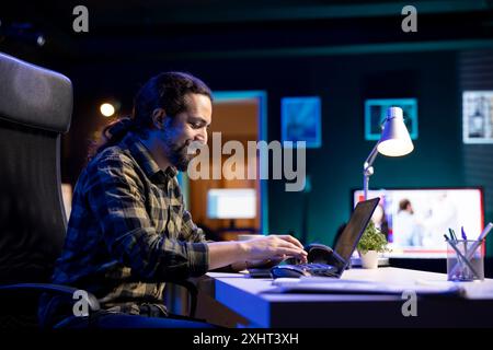 Smiling man at his living room desk, typing and browsing the internet on a laptop. Positive guy is concentrating on finishing several tasks as he sends emails on his electronic device. Stock Photo