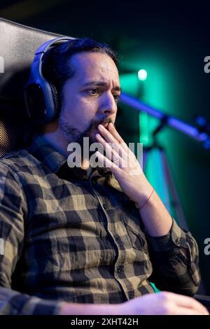 Shocked man sitting at a desk, watching a movie on portable computer while wearing headphones. Young guy listening to a podcast on wireless headset while surfing the net on laptop. Stock Photo