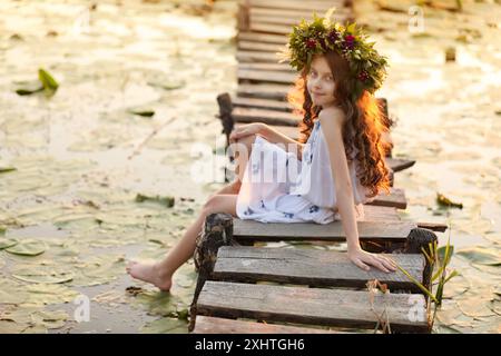 Cute little girl wearing wreath made of beautiful flowers on pier near pond Stock Photo