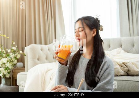 An attractive Asian woman is keeping her eyes closed while drinking orange juice at a coffee table in the living room. people, food, lifestyle, domest Stock Photo