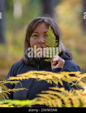 Close-up portrait of a smiling woman 50 years old holding a fern leaf to her face in an autumn forest. Stock Photo