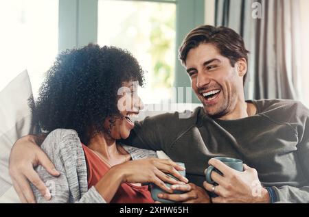 Couple, happy and drinking coffee with laughing, relaxing and bonding in lounge, sofa and home on weekend break. Woman, man and interracial partner Stock Photo