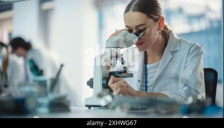 Portrait of Young Professional Female Engineer Using a Microscope in a Lab to Examine an Electrical Component. Successful Woman Working as Laboratory Specialist in an Industrial Manufacturing Company Stock Photo