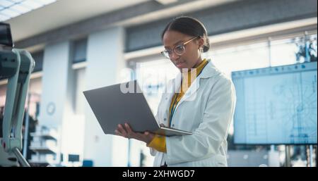 Portrait of Young Black Female Specialist in Lab Coat Using Laptop to Test an AI Robotic Prototype. Professional, Successful Woman Working as an Engineer in Modern High Tech Company Startup Stock Photo