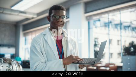 Portrait of Black Young Male Scientist Using Laptop Computer, Looking at Camera and Smiling in a Laboratory. Professional African American Man Working as Engineer, Monitoring Technology Projects Stock Photo