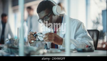 Portrait of Black Male Lab Specialist Soldering a Circuit Board, Using Headphones in an Industrial Company. Professional Man Listening to Music, Working on Technological Project For a Startup Stock Photo