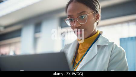 Portrait of Young Black Female Expert in a Lab Coat Smiling and Using Laptop Computer. Professional and Successful Woman Working as an Engineer, Developing High Technology Projects Stock Photo