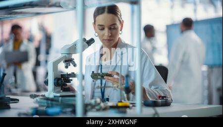 Diverse Young Group of Engineers Working in a Startup Lab. Female Specialist Checking on Electronic Circuit Using Microscope While Project Managers Discuss Manufacturing Using Laptop Computer Stock Photo