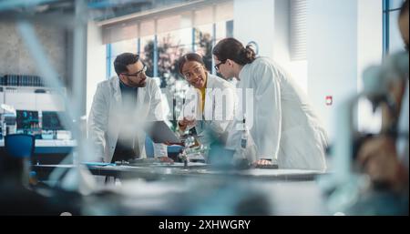 Diverse Young Group of Engineers Working in a Startup Company Lab. Project Managers Discuss Manufacturing Using Laptop Computer While Female Expert Checking on Electronic Circuit Using Microscope Stock Photo