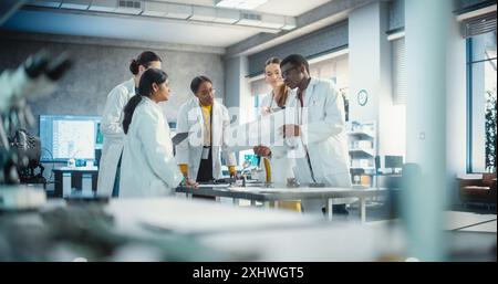 Diverse Team of Industrial Robotics Specialists Gathered Around a Table to Discuss a Blueprint. Engineers In Lab Coats Using Laptop Computer and Tablet, Brainstorming Ideas For Electronic Components Stock Photo