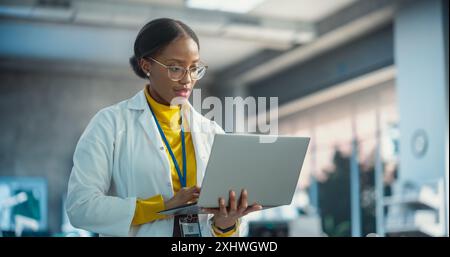 Portrait of Young Black Female Specialist in a Lab Coat Using Laptop. Professional and Successful Woman Working as an Engineer, Thinking and Developing High Technology Projects In Startup Stock Photo