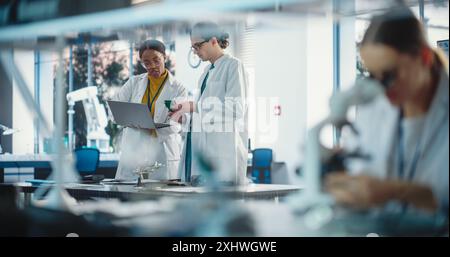 Diverse Young Group of Engineers Working in a Startup Lab. Two Project Managers Discuss Manufacturing Using Laptop Computer While Female Specialist Checks on Electronic Circuit Using Microscope Stock Photo
