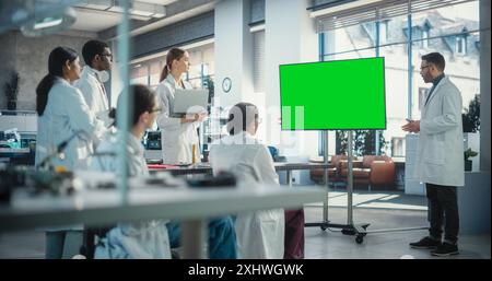 Group of Students Listening to Their Robotics Professor Who Uses Green Screen Monitor in a Laboratory. Team of Engineers Having a Meeting and Discussing Manufacturing Ideas, Using Technology Stock Photo