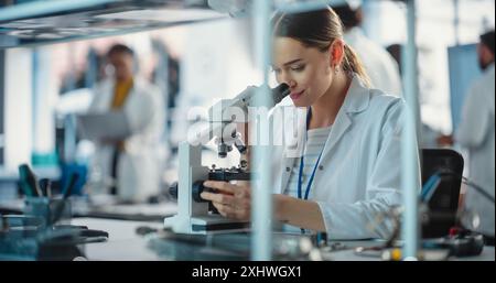 Diverse Young Group of Engineers Working in a Startup Lab. Project Managers Discuss Manufacturing Using Laptop Computer While Female Specialist Checking on Electronic Circuit Using Microscope Stock Photo