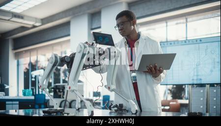 Young African American Male Engineer Testing Industrial Programmable Robot Animal in a Factory Development Workshop. Black Researcher in a Lab Coat Developing AI Pet Prototype, Using Laptop Stock Photo