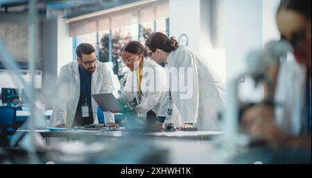 Diverse Young Group of Engineers Working in a Startup Company Lab. Project Managers Discuss Manufacturing Using Laptop Computer While Female Specialist Checking on Electronic Circuit Using Microscope Stock Photo