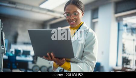 Portrait of Young Black Female Specialist in a Lab Coat Smiling and Using Laptop Computer. Professional and Successful Woman Working as an Engineer, Developing High Technology Projects Stock Photo