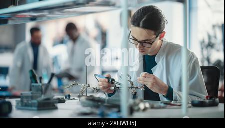 Diverse Young Group of Engineers Working in a Startup Lab. Male Specialist Soldering a Circuit Board While Team of Professionals Discuss Technological Project Strategy Using Laptop Computer. Stock Photo