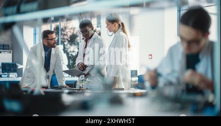Diverse Young Group of Engineers Working in a Modern Startup Lab. Male Specialist Soldering a Circuit Board While Team of Professionals Discuss Technological Project Strategy Using Laptop Computer. Stock Photo