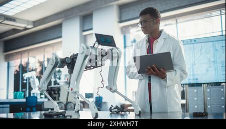 Young African American Male Engineer Testing Industrial Programmable Robot Animal in a Factory Development Workshop. Black Researcher in a Lab Coat Developing AI Canine Prototype, Using Laptop Stock Photo