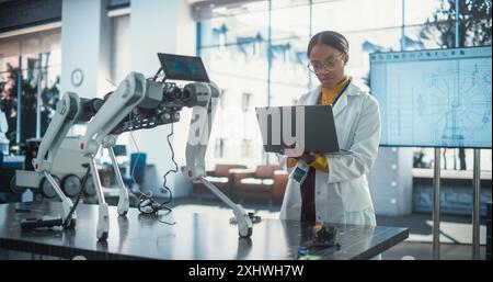 Portrait of Young Black Female Specialist in Lab Coat Using Laptop to Test an AI Robotic Prototype. Professional, Successful Woman Working as an Engineer in Modern High Technology Company Startup Stock Photo