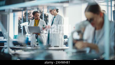 Diverse Young Group of Engineers Working in a Startup Lab. Two Project Managers Discuss Manufacturing Using Laptop Computer While Female Specialist Checking on Electronic Circuit Using Microscope Stock Photo