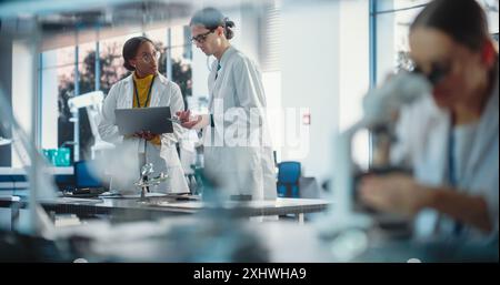 Diverse Young Group of Engineers Working in a Startup Lab. Female Specialist Checking on Electronic Circuit Using Microscope While Two Project Managers Discuss Manufacturing Using Laptop Computer Stock Photo