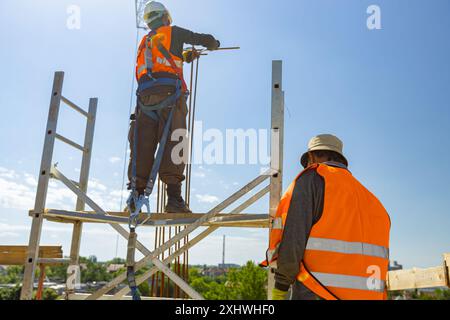 Supervisor, foreman look out on worker tying armature to make a reinforcing frame for concrete pillar, he carry fall arrestor device, hooks for safety Stock Photo