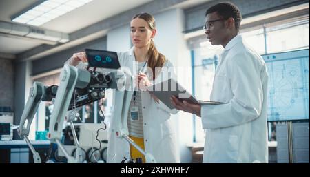 Two Multiethnic Industrial Robotics Specialists Working on a Mobile Robot, Using Laptop. Black Male Young Engineer In Lab Coat Discussing an Automated AI Robotic Dog with his Female Project Supervisor Stock Photo