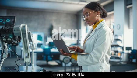 Portrait of Young Black Female Specialist in Lab Coat Using Laptop to Test an AI Robotic Prototype. Professional, Successful Woman Working as an Engineer in High Technology Company Startup Stock Photo