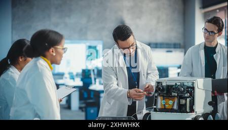 Diverse Team of Industrial Robotics Specialists Gathered Around a Table With Mobile Robot. Engineers In Lab Coats Discussing an Automated AI Robotic Delivery Assistant, Using Laptop and Tablet Stock Photo