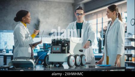 Team of Industrial Specialists Working on Mobile Robot, Using Laptop. Young Male and Female Engineers in a Lab Discussing Automated AI Robotic Delivery Assistant, Brainstorming Improvement Ideas Stock Photo