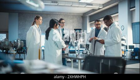 Diverse Team of Industrial Robotics Specialists Gathered Around a Table With a Mobile Robot. Engineers In Lab Coats Discussing an Automated AI Robotic Dog, Using Laptop and Tablet, Brainstorming Stock Photo