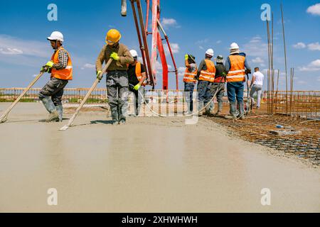 Team of construction workers are pouring and leveling fresh concrete using handmade tool over rectangular armature. Stock Photo