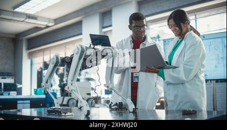 Male Project Manager and Female Machinery Operator Collaborate on a Mobile Robot Animal Project, Using Laptop Computer. Two Multiethnic Engineers Brainstorming, Discussing Futuristic AI Pet Stock Photo