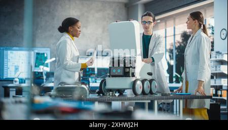 Team of Industrial Specialists Working on Mobile Robot, Using a Laptop. Young Male and Female Engineers in a Lab Discussing an Automated AI Robotic Delivery Assistant, Brainstorming New Ideas Stock Photo